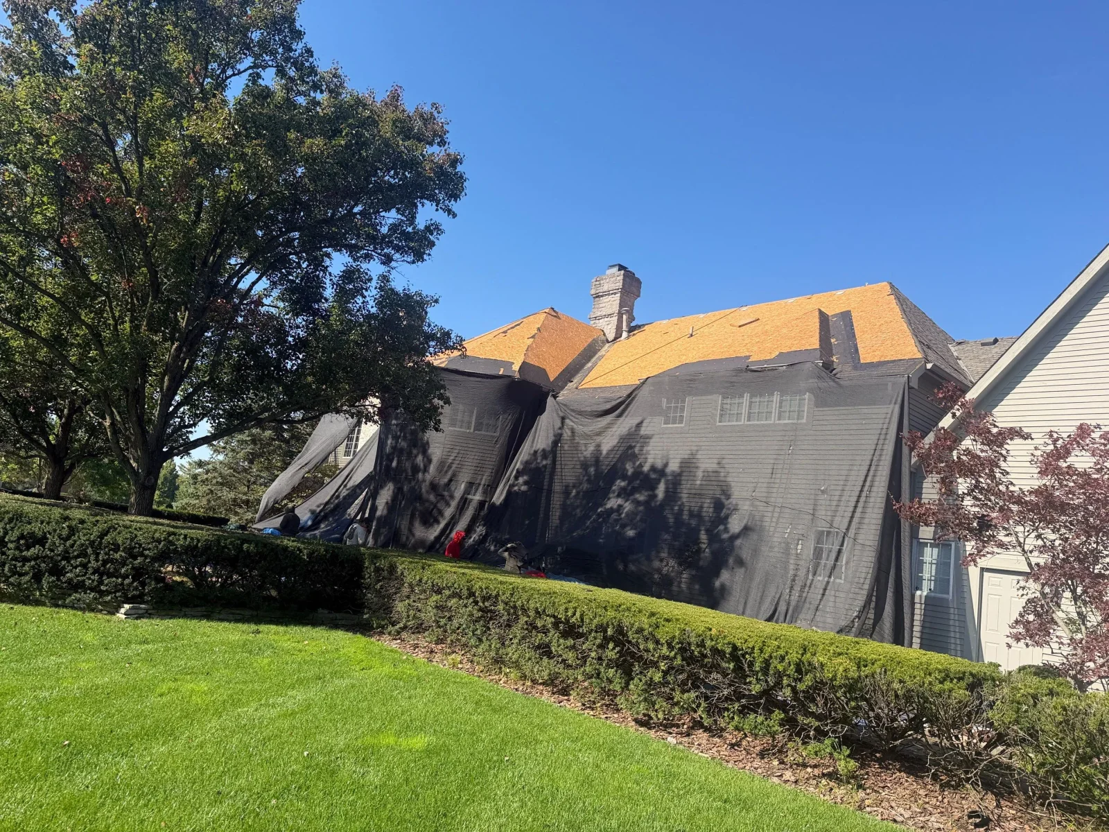 Roof tear-off in progress on a two-story home showing exposed roof decking and black debris netting over siding