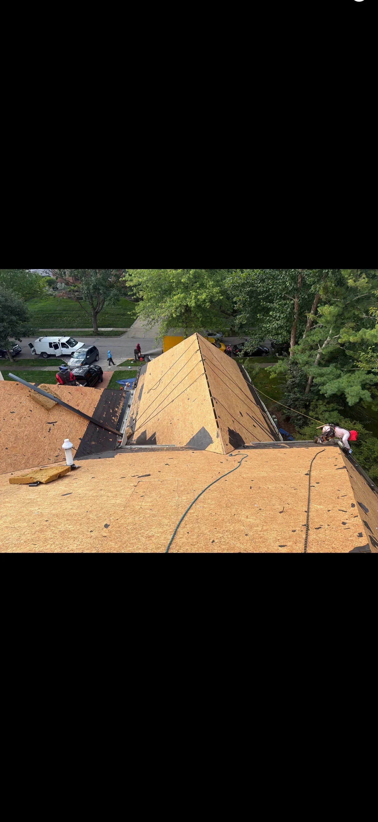 Pyramid roof with new OSB sheathing during tear-off, old black underlayment and debris on lower slopes, worker in harness at eave
