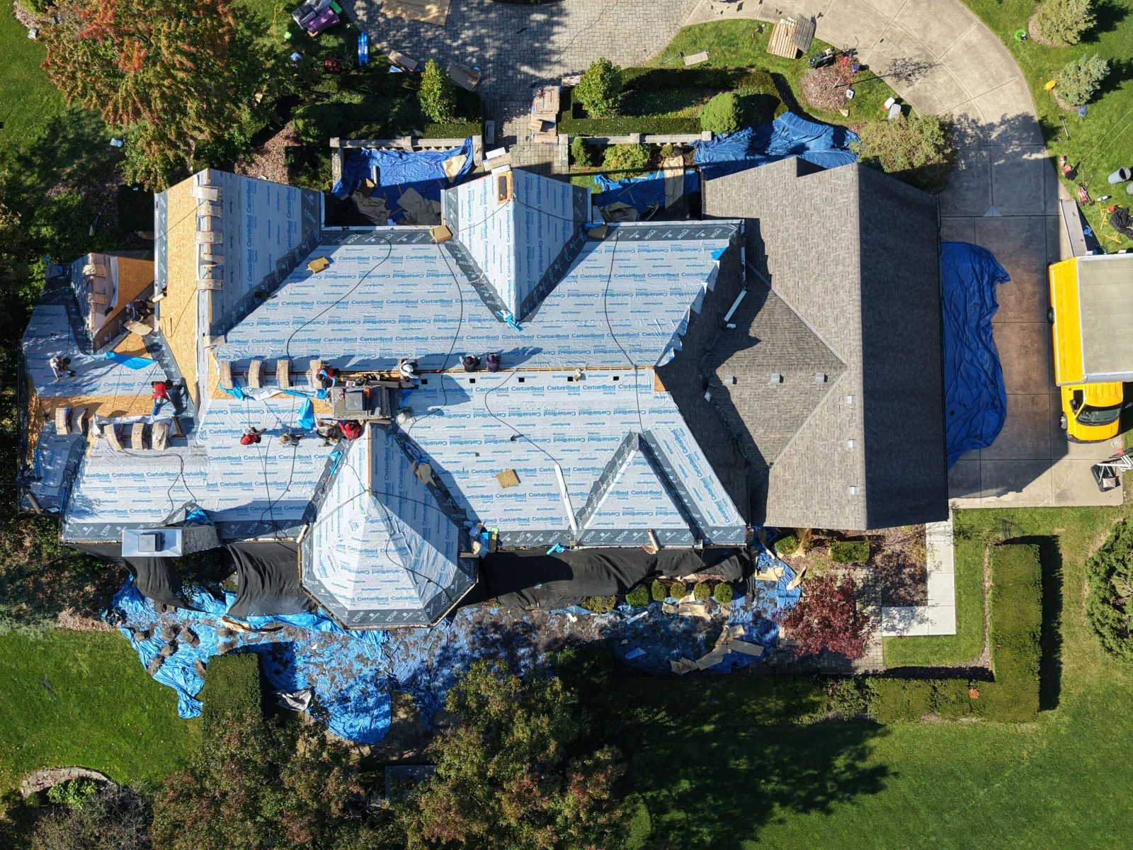 Aerial view of roof replacement with CertainTeed synthetic underlayment and exposed plywood decking, blue tarps, and crew working on steep roof sections