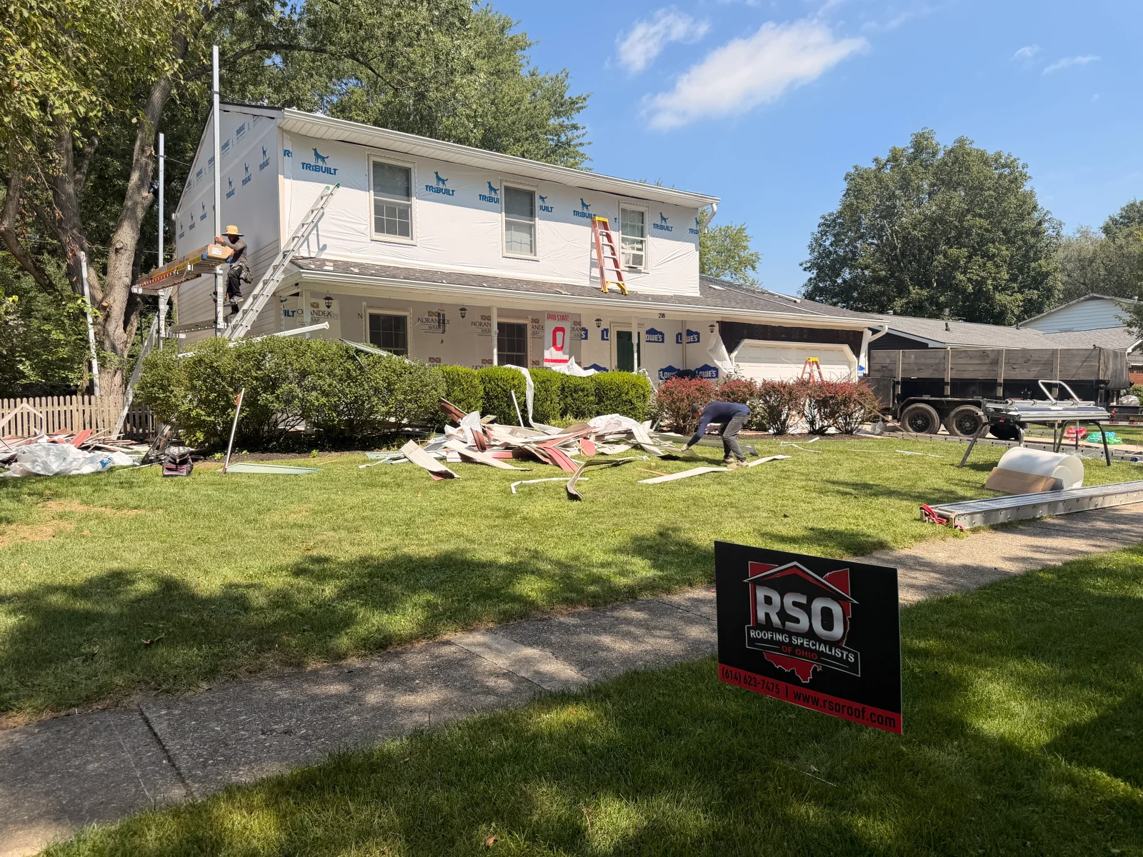 Two-story home with roof being torn off, shingle debris on lawn, house wrap on walls, and dump trailer in driveway