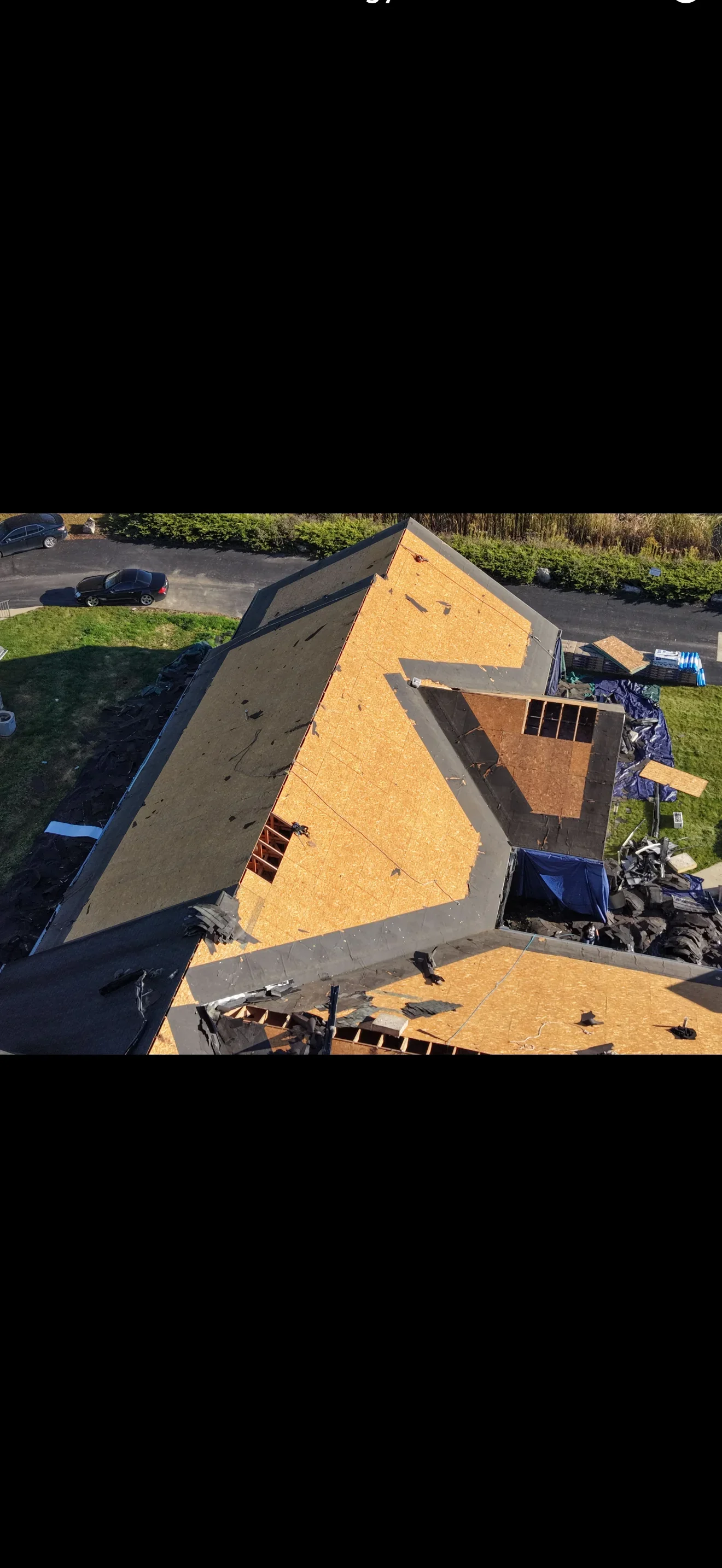 Aerial view of a steep residential roof stripped to OSB sheathing with exposed framing, dormer opening, and remaining black underlayment during tear-off