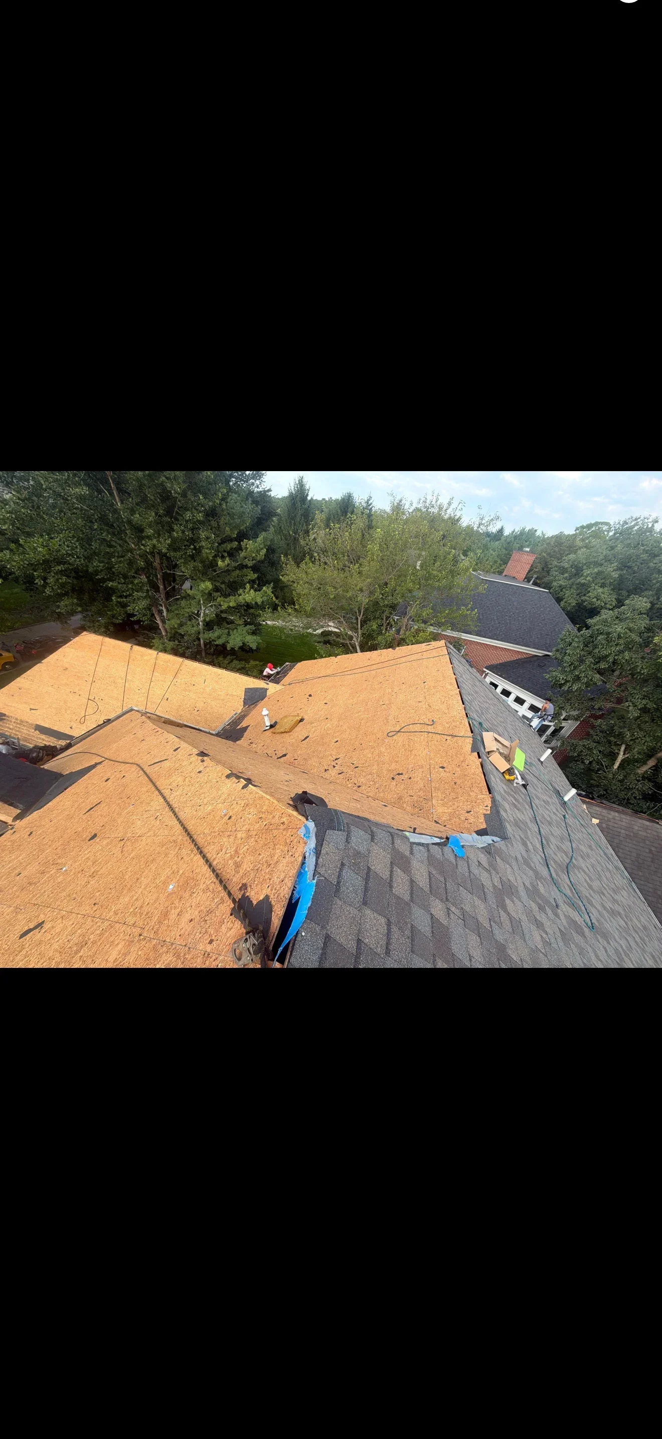 Residential roof with exposed OSB sheathing during tear-off, asphalt shingles and blue tarp at roof transition