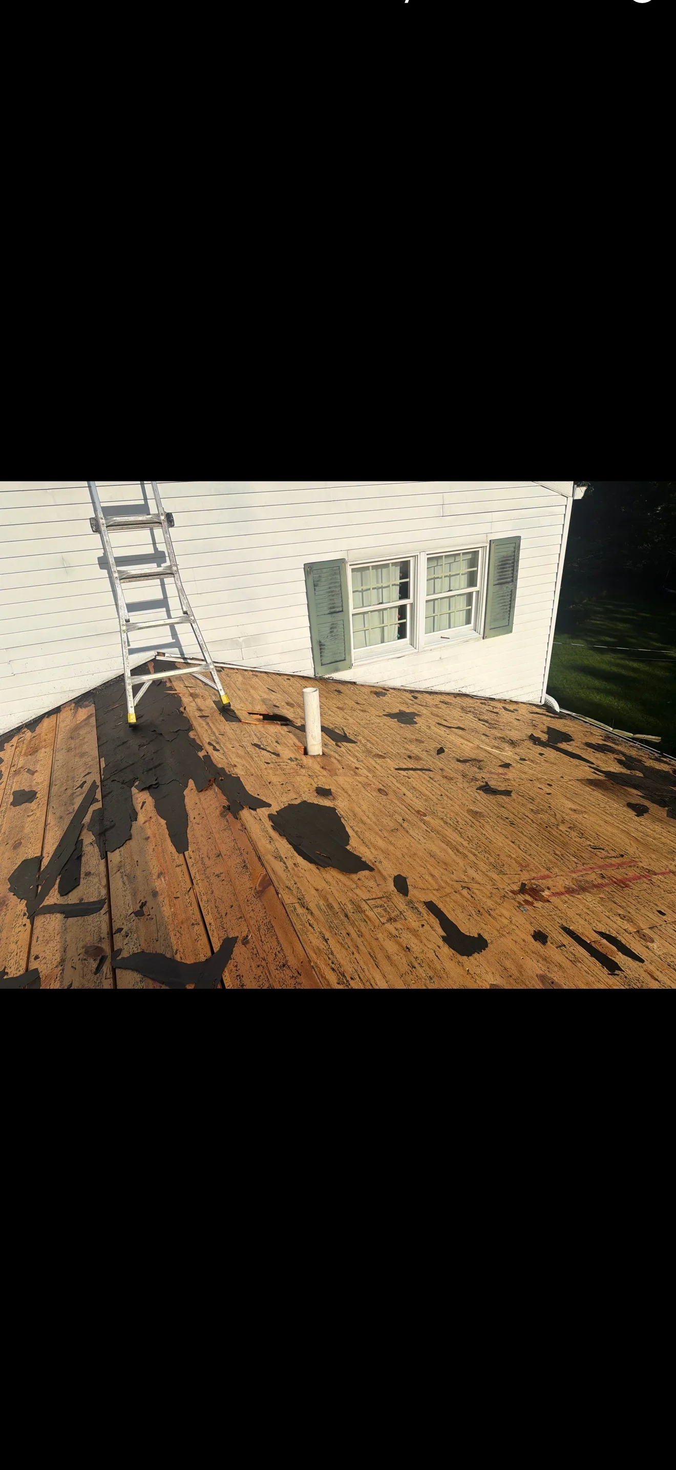 Bare plywood roof deck with remnants of black roofing felt, white PVC vent pipe, and aluminum ladder during tear-off