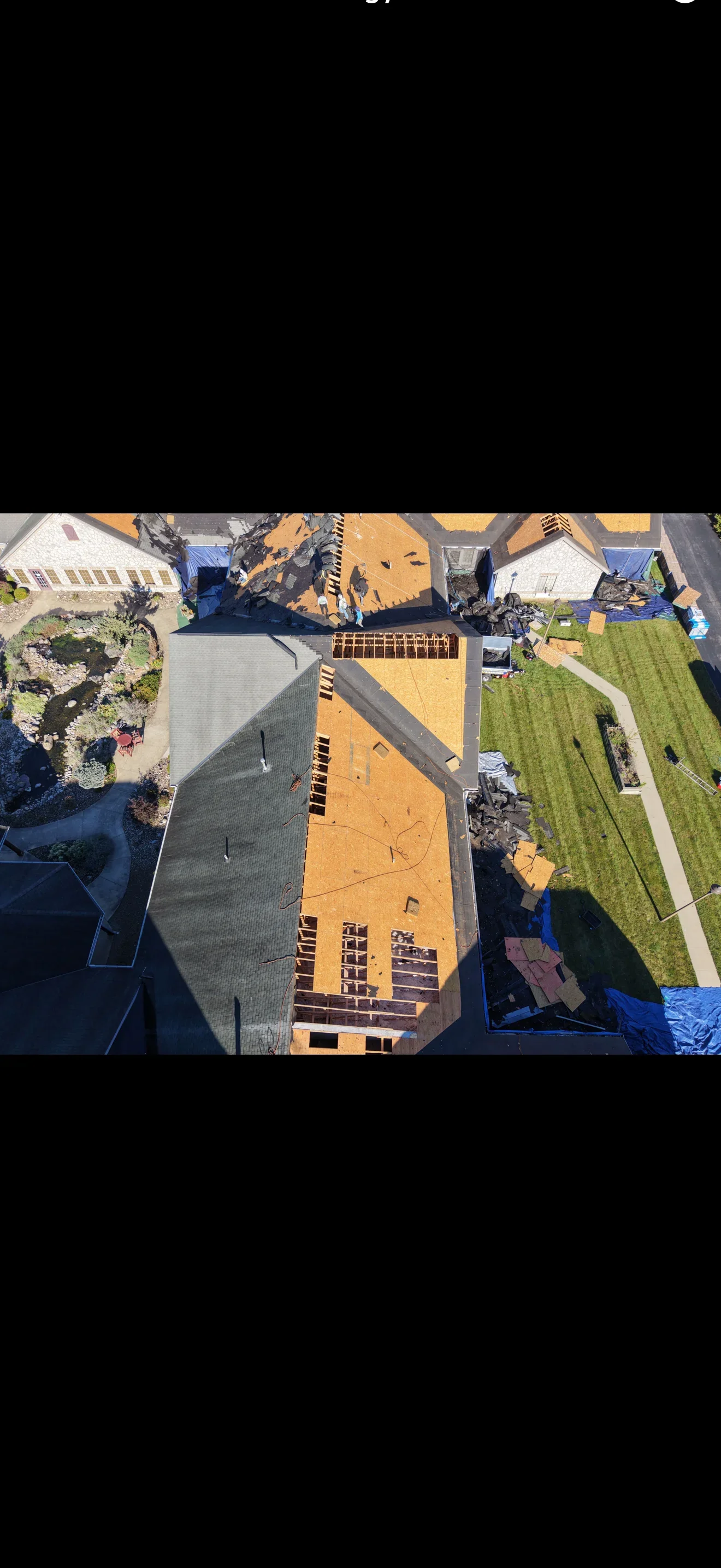 Aerial view of residential roof tear-off with exposed OSB decking, wood framing, and blue tarps over asphalt shingle debris
