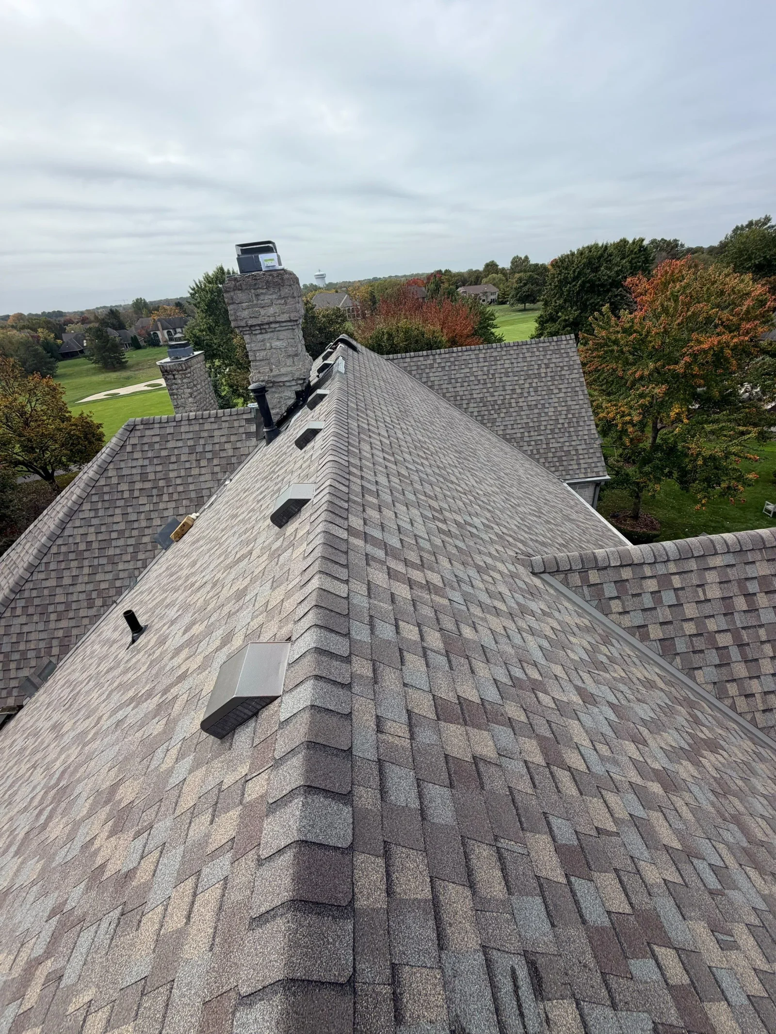 Gray and tan architectural asphalt shingle roof with box vents, plumbing vent stack, and stone chimney