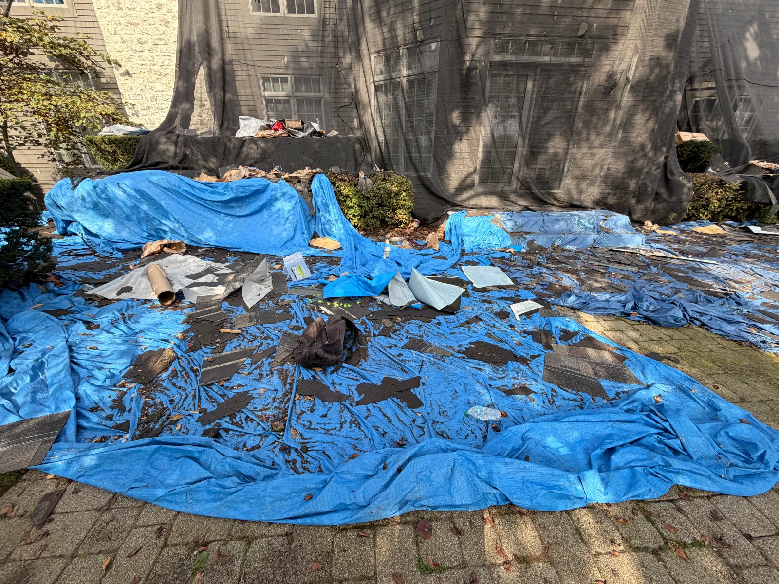 Blue tarp covered with asphalt shingle debris and black debris netting on a home exterior during roof tear-off