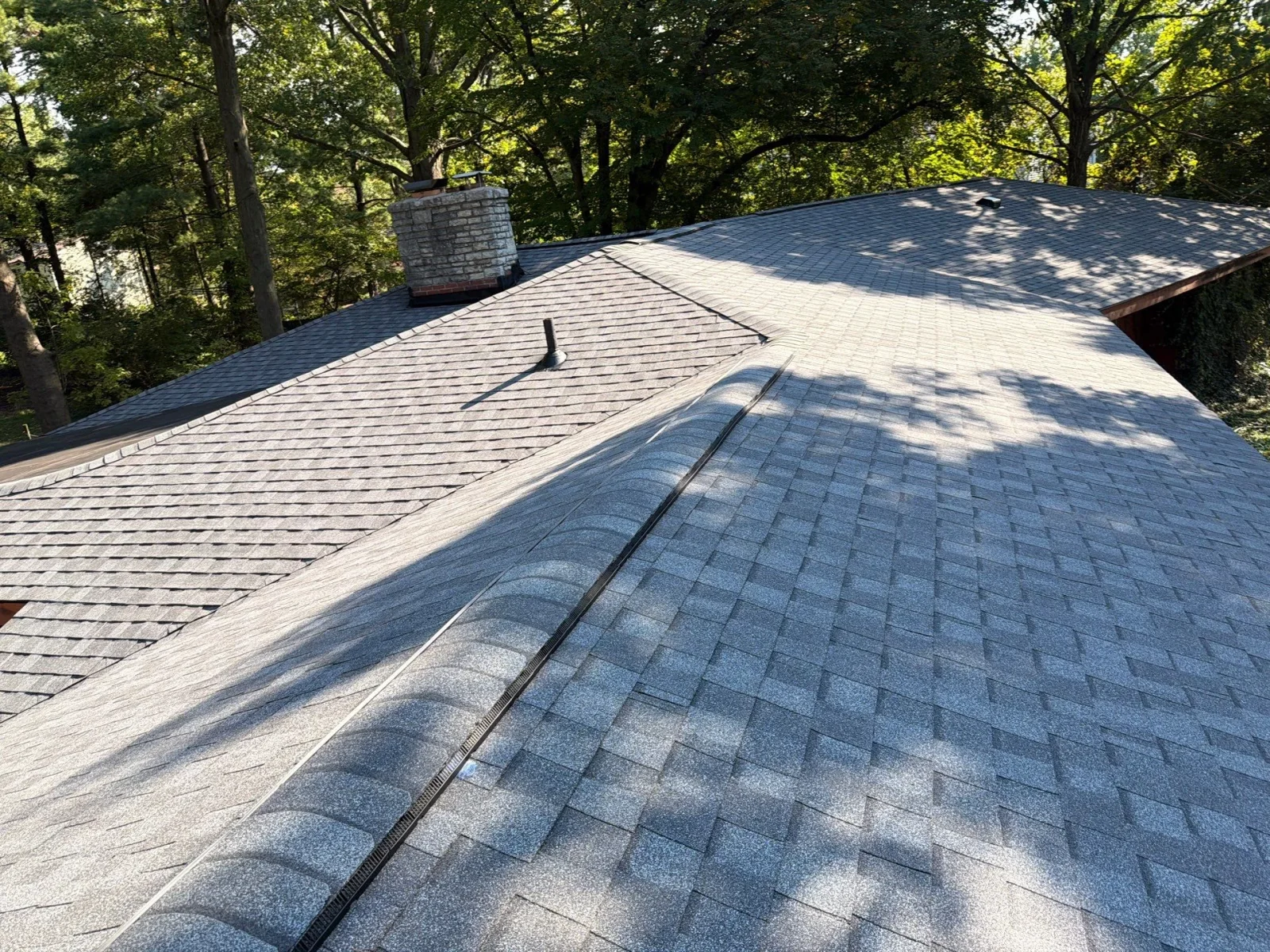 Gray asphalt shingle roof with black ridge vent, stone chimney, and pipe vent boot