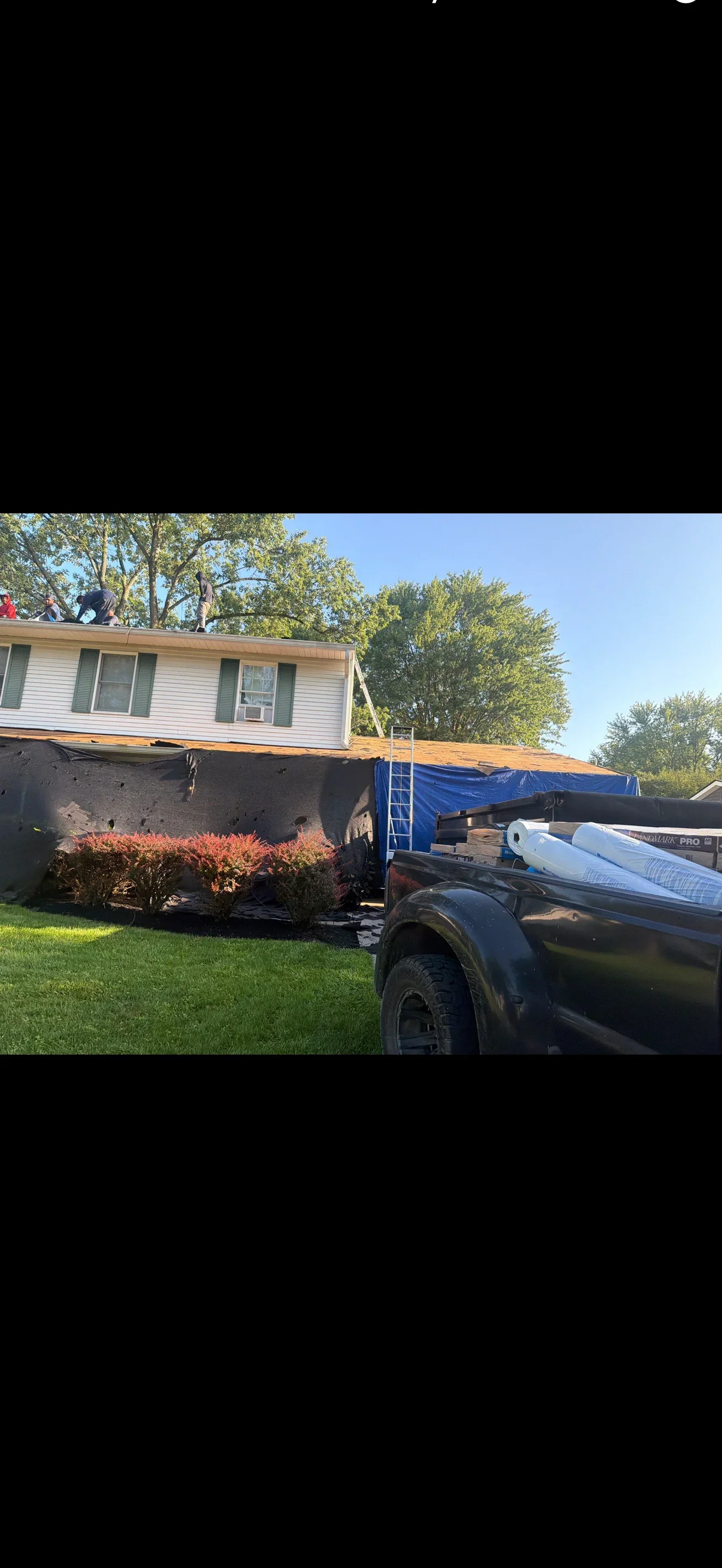 Crew working on a roof tear-off with exposed OSB sheathing, black underlayment, blue tarp, and Landmark PRO shingle bundles in a truck bed