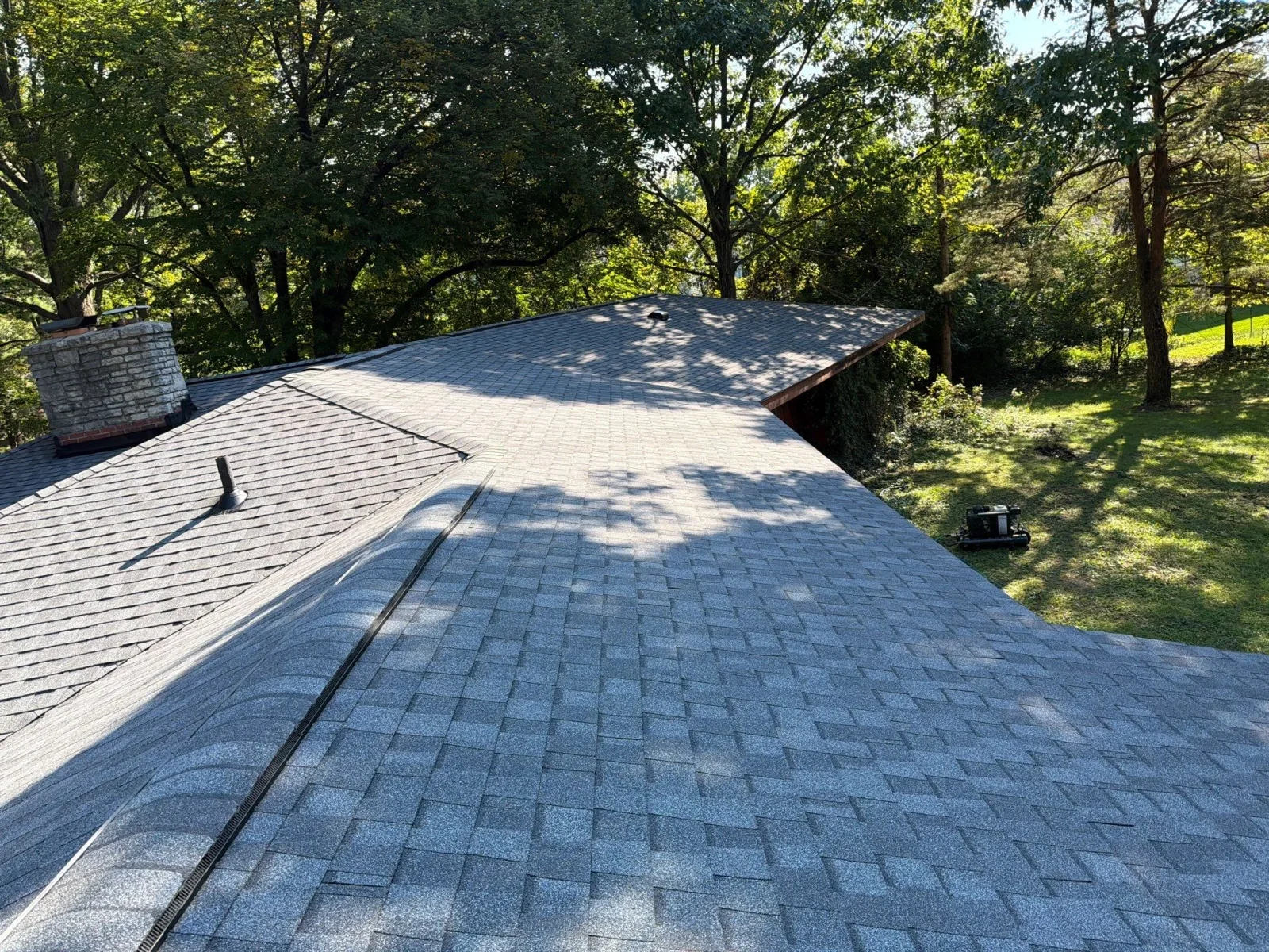 Blue-gray architectural asphalt shingle roof with metal valley flashing, stone chimney, and black plumbing vent pipe