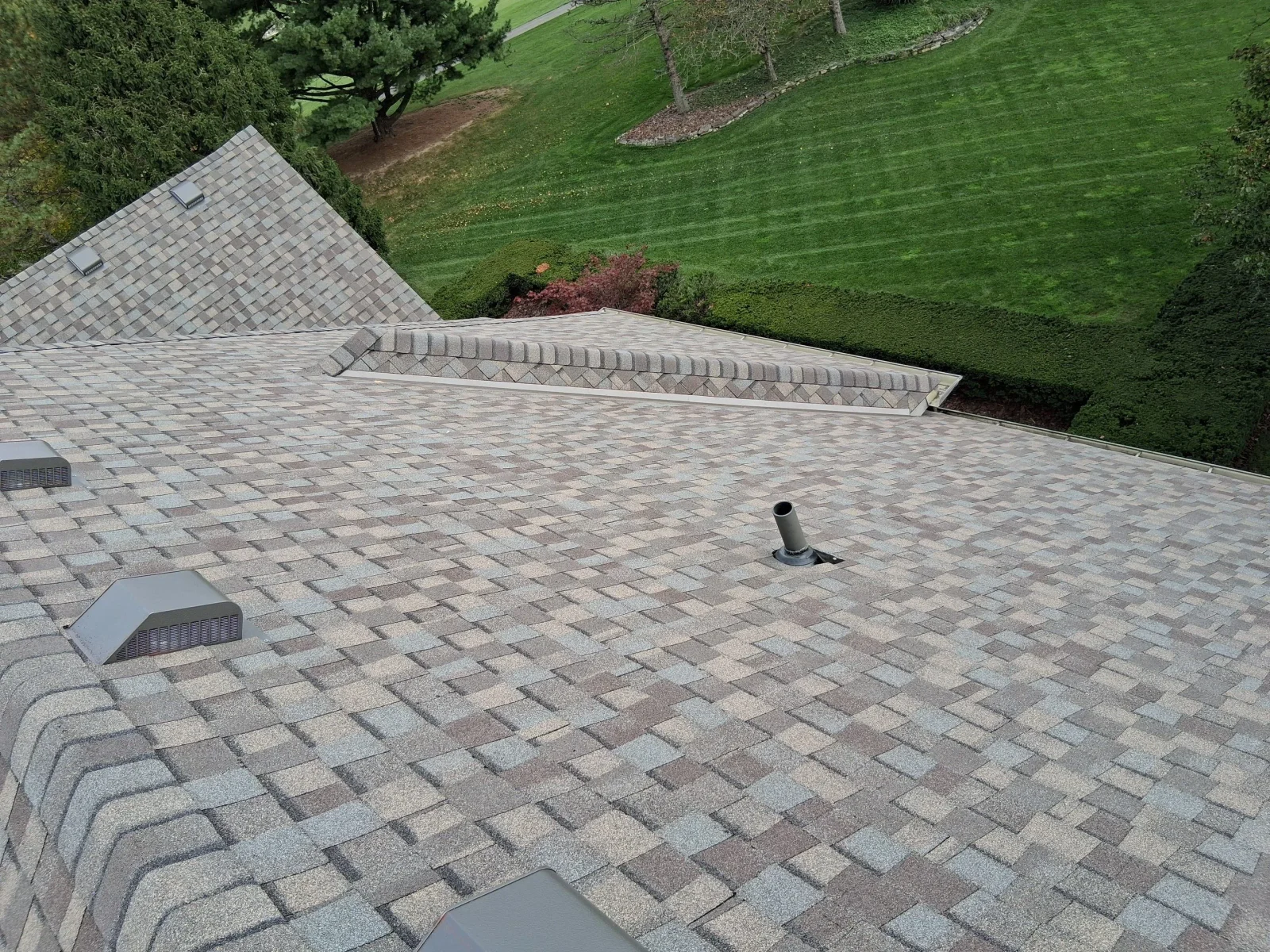 Architectural asphalt shingle roof in tan, gray, and mauve tones with gray box vents, plumbing vent pipe, and ridge cap