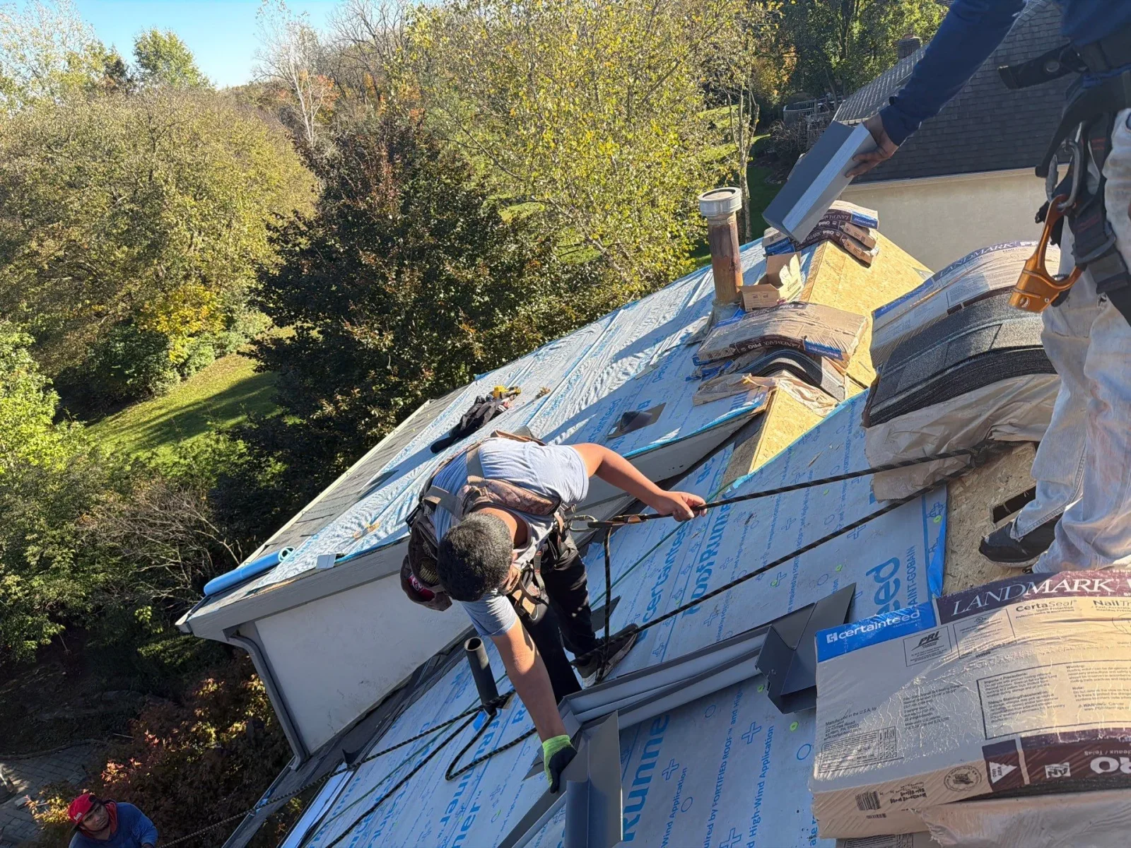 Roofer in fall protection harness installing blue synthetic underlayment and staging Landmark shingles on a steep roof