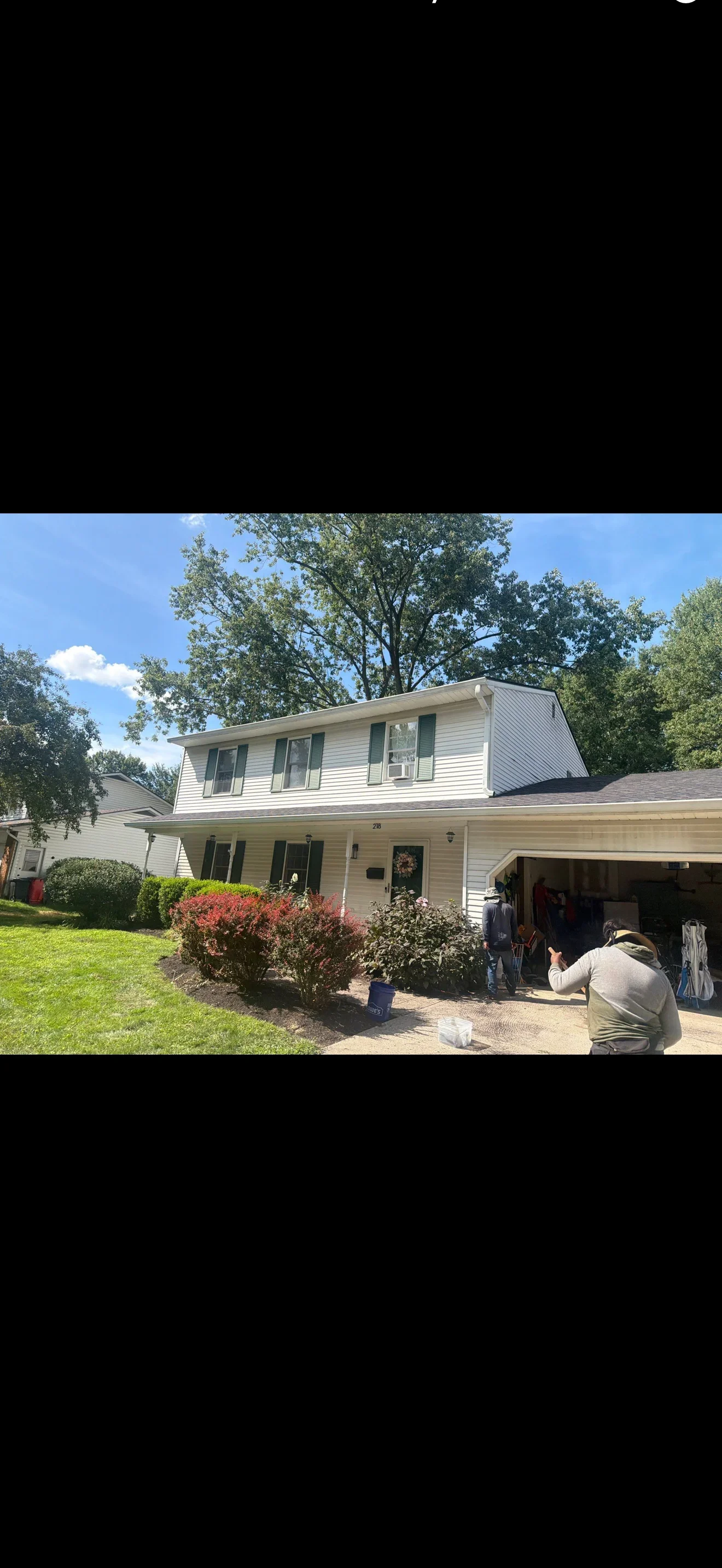 Two-story home with white vinyl siding, green shutters, dark asphalt shingle roof, and white gutters with crew working in driveway