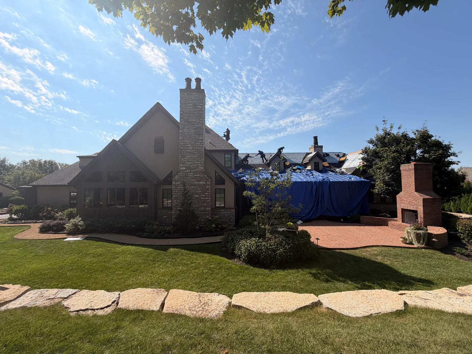 Large home with stucco walls and stone chimney, roof partially covered by blue tarps while workers replace roofing on steep gabled planes